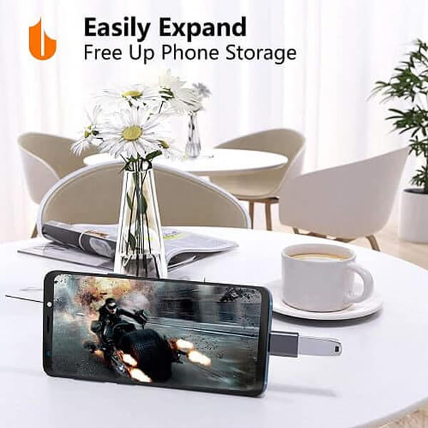 A smartphone connected to a Generic USB Type-C to OTG Adapter (USB 3.1 Male to USB 3.0 Female Converter for MacBook Pro, Air & iPad Pro in Black) is placed on a table with a cup, a book, and flowers. The screen shows a motorcycle scene with the text: "Easily Expand. Free Up Phone Storage with High-Speed Data Transfer.