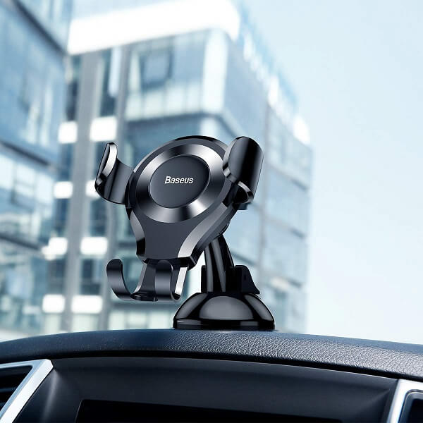 A BASEUS Gravity Universal Car Phone Holder with Adhesive Mount is attached to a car dashboard, allowing hands-free phone use, with a modern glass building in the background.
