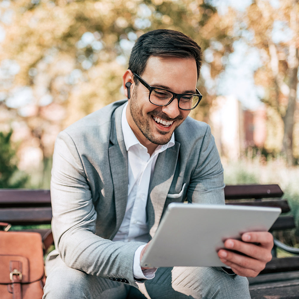 A man in a gray suit sits on an outdoor bench, smiling at his tablet. He wears glasses and black JAM-AUDIO Jam True Wireless In-Ear - Executive Headphones for hands-free calling, enjoying true wireless freedom with his brown bag beside him.