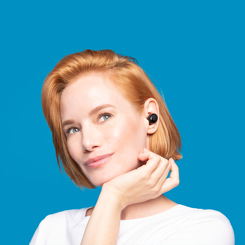 A woman with short red hair wears a white shirt and the JAM-AUDIO Jam Audio Live Loud TWS BT Headset in black, highlighting hands-free calling as she poses with her hand under her chin against a blue background.