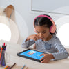 A young girl wearing BELKIN SoundForm Mini Wireless On-Ear Headphones for Kids in Pink with volume limitation using a tablet at a desk with colored pencils in the background for distance learning.