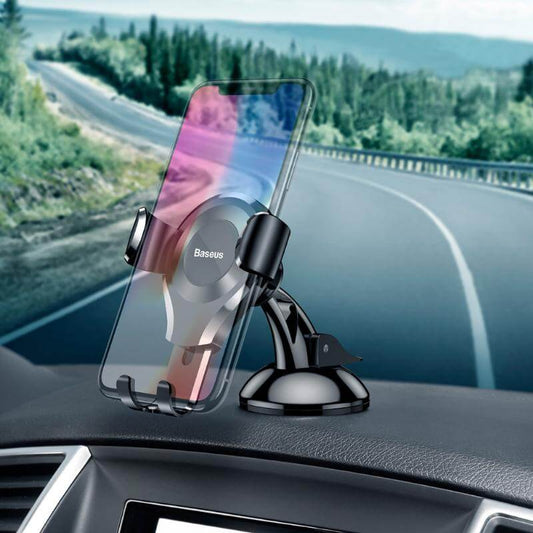 A smartphone mounted on a BASEUS Gravity Universal Car Phone Holder for Windshield and Dashboard in a car, with a curving road and forested landscape visible through the windshield. The adjustable mount from Baseus features a retractable gravity mechanism for effortless phone security and stability.